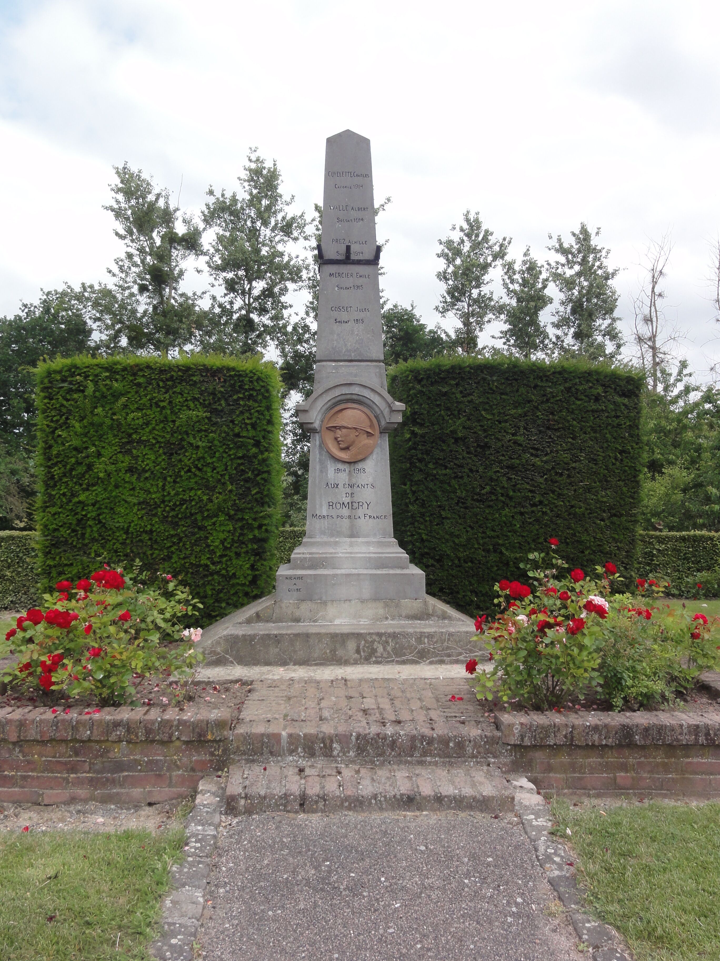 A memorial monument with names and dates, surrounded by hedges and red roses.