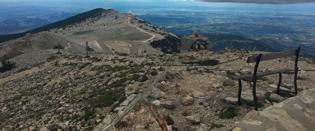 A mountainous landscape with a rocky path, a wooden bench, and a distant view of a valley under a cloudy sky.