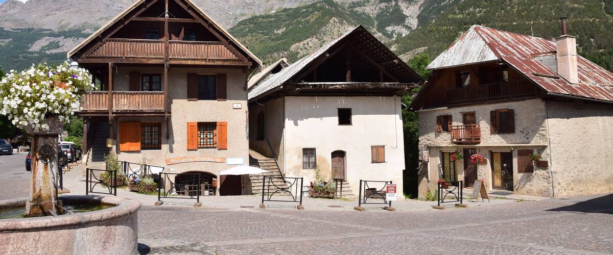 A cobblestone square with traditional wooden buildings, a fountain, and mountains in the background.