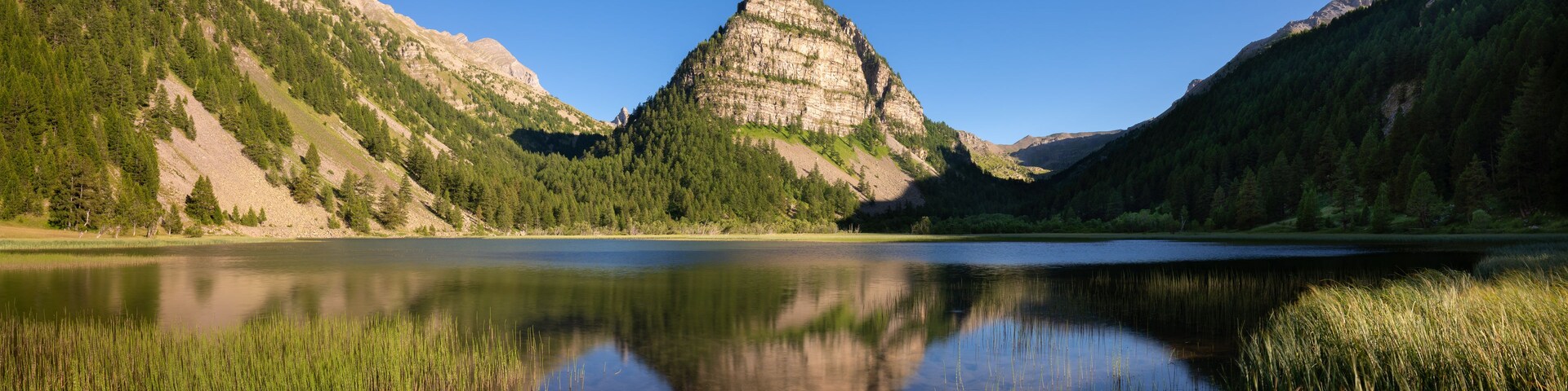 Un lac de montagne aux eaux limpides, dont le miroir reflète le paysage, entouré de collines boisées.