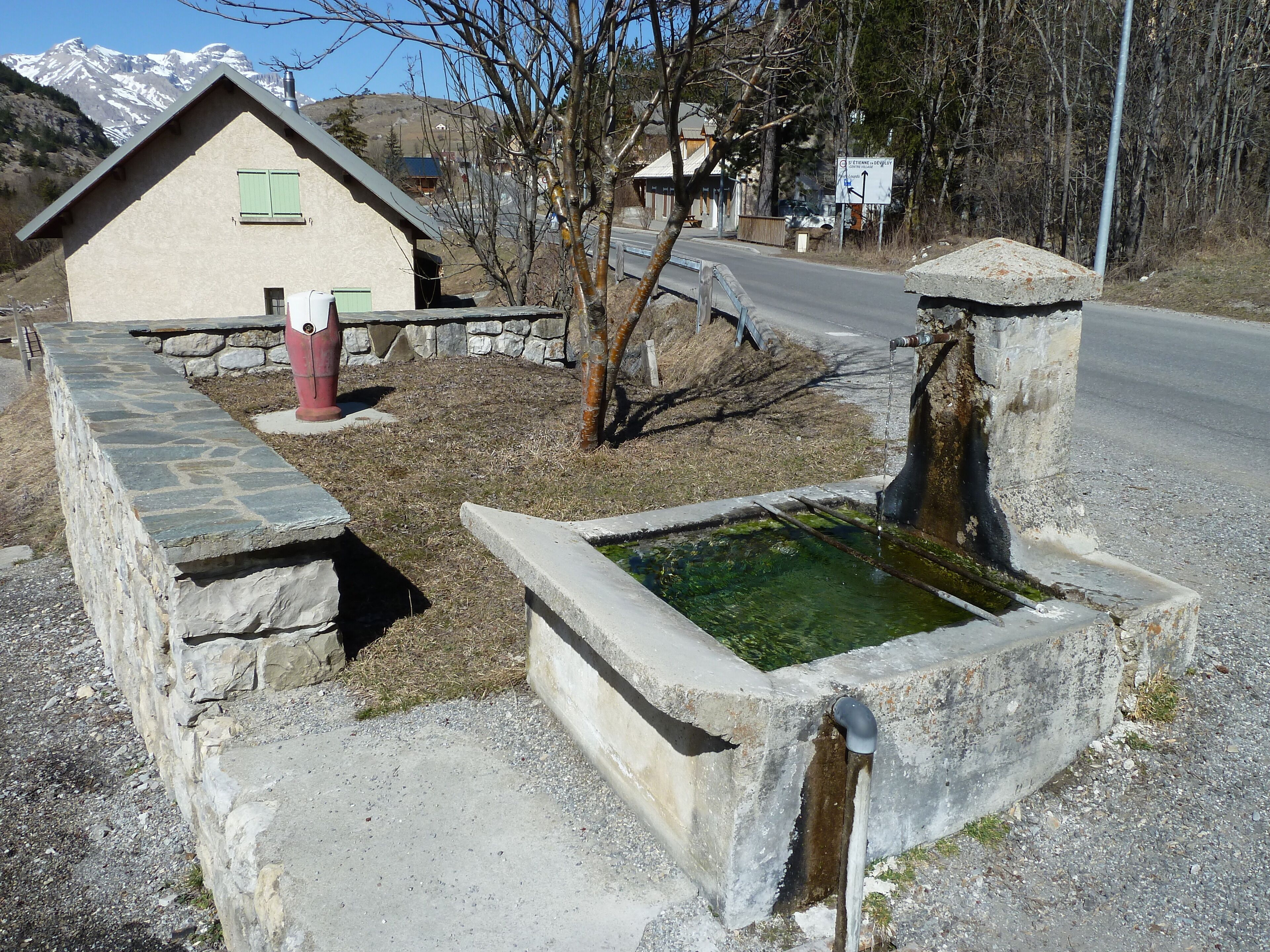 A stone well with water, a stone wall, a small house with green shutters, and a mountain in the background.