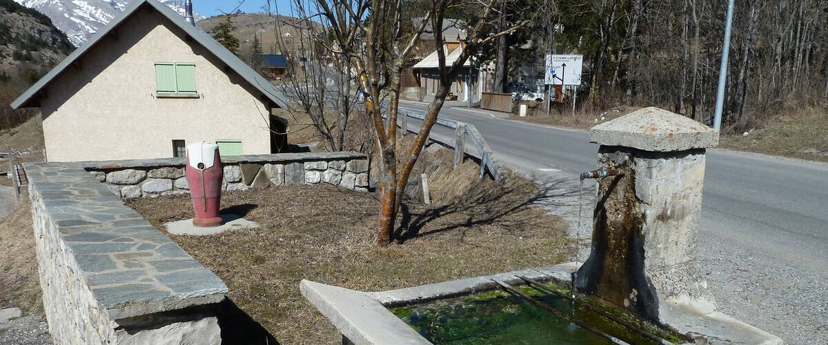 A stone well with water, a stone wall, a small house with green shutters, and a mountain in the background.