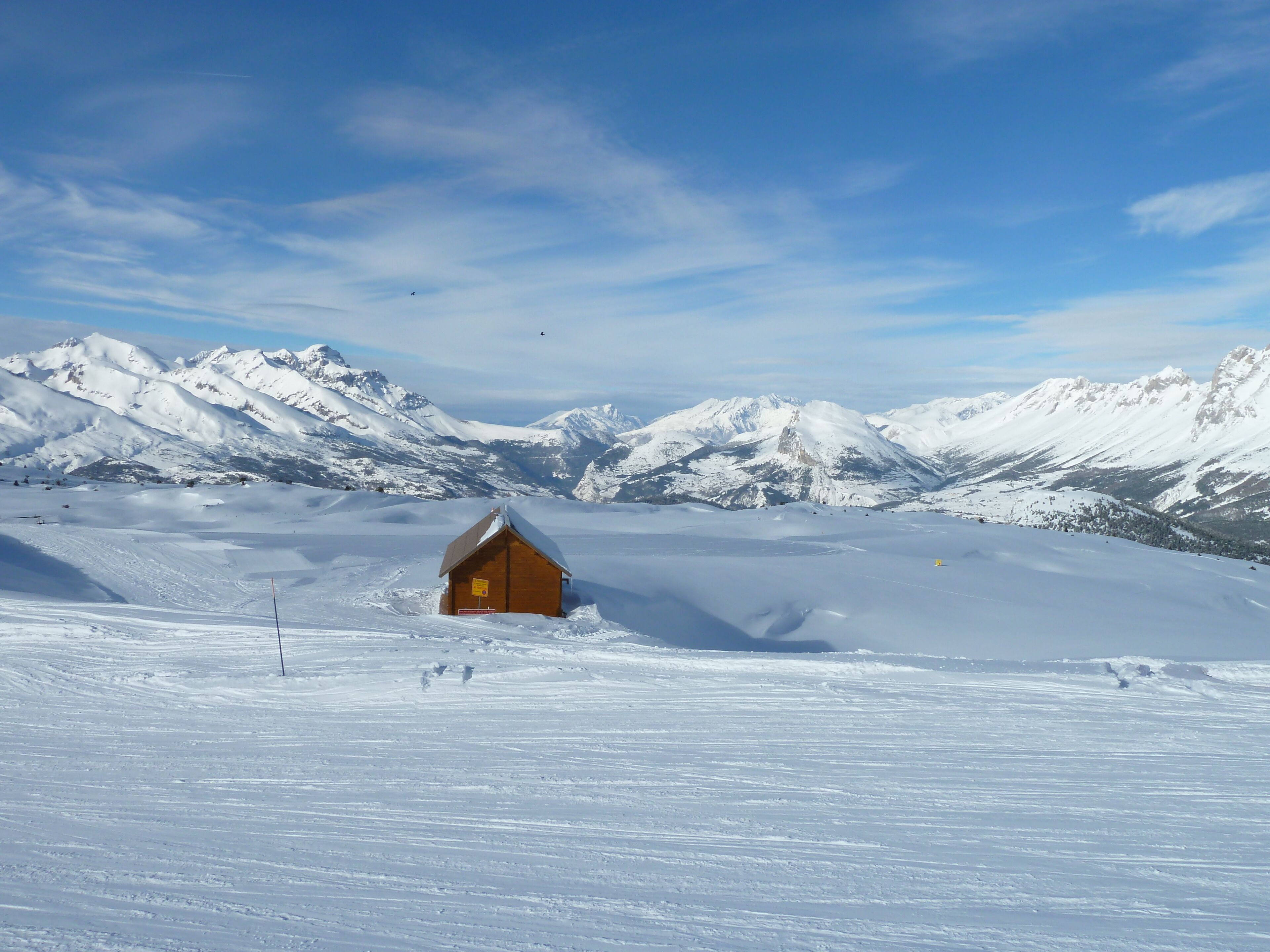 A wooden cabin in a snowy mountain landscape with snow-covered peaks in the background.