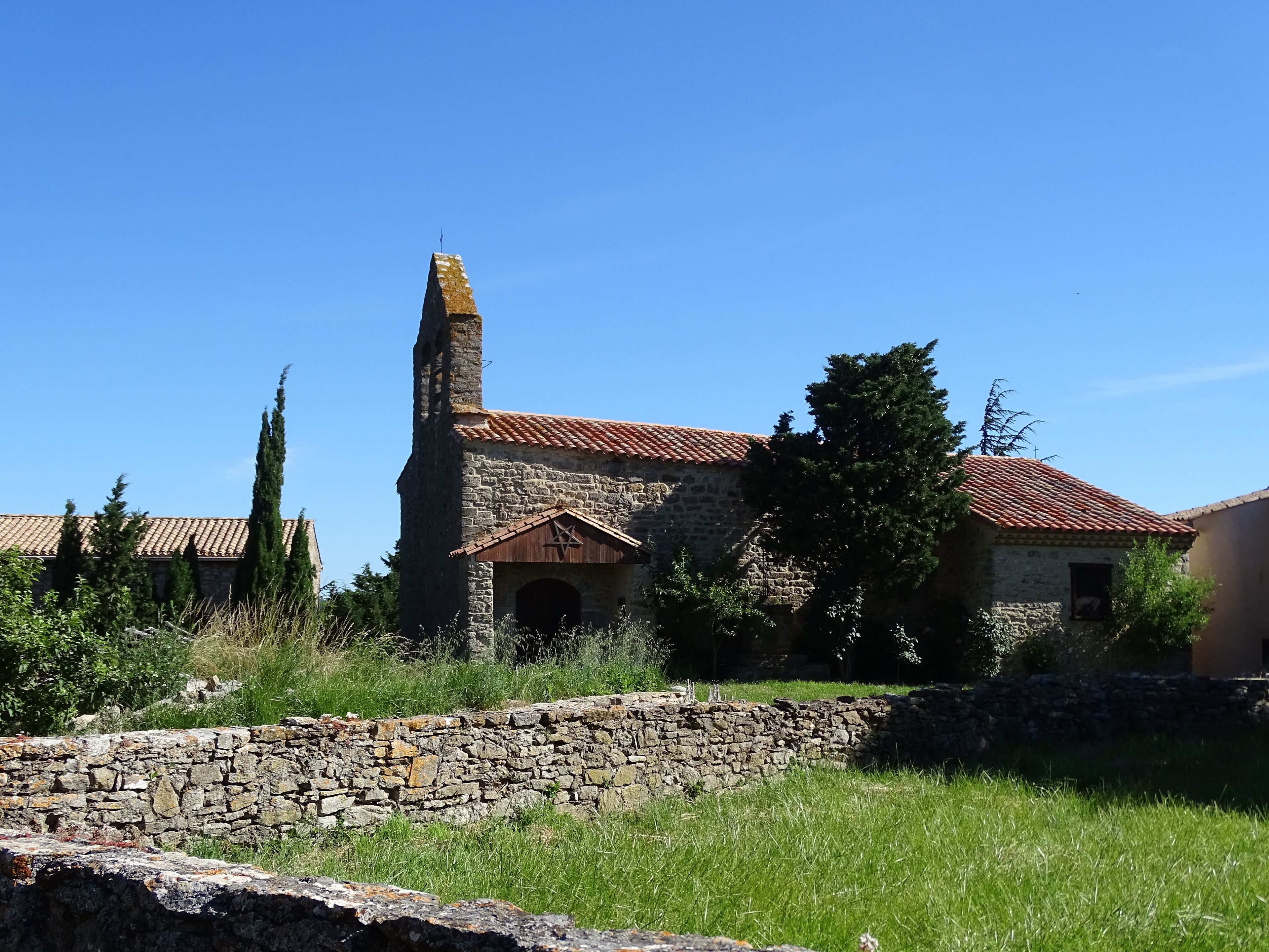An old stone building with a red-tiled roof, surrounded by greenery and a stone wall.