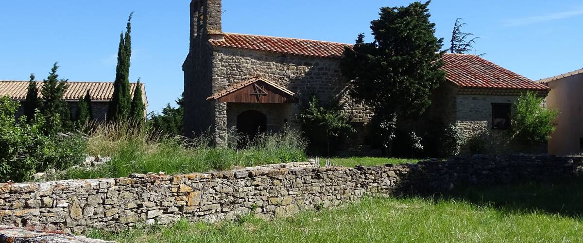 An old stone building with a red-tiled roof, surrounded by greenery and a stone wall.