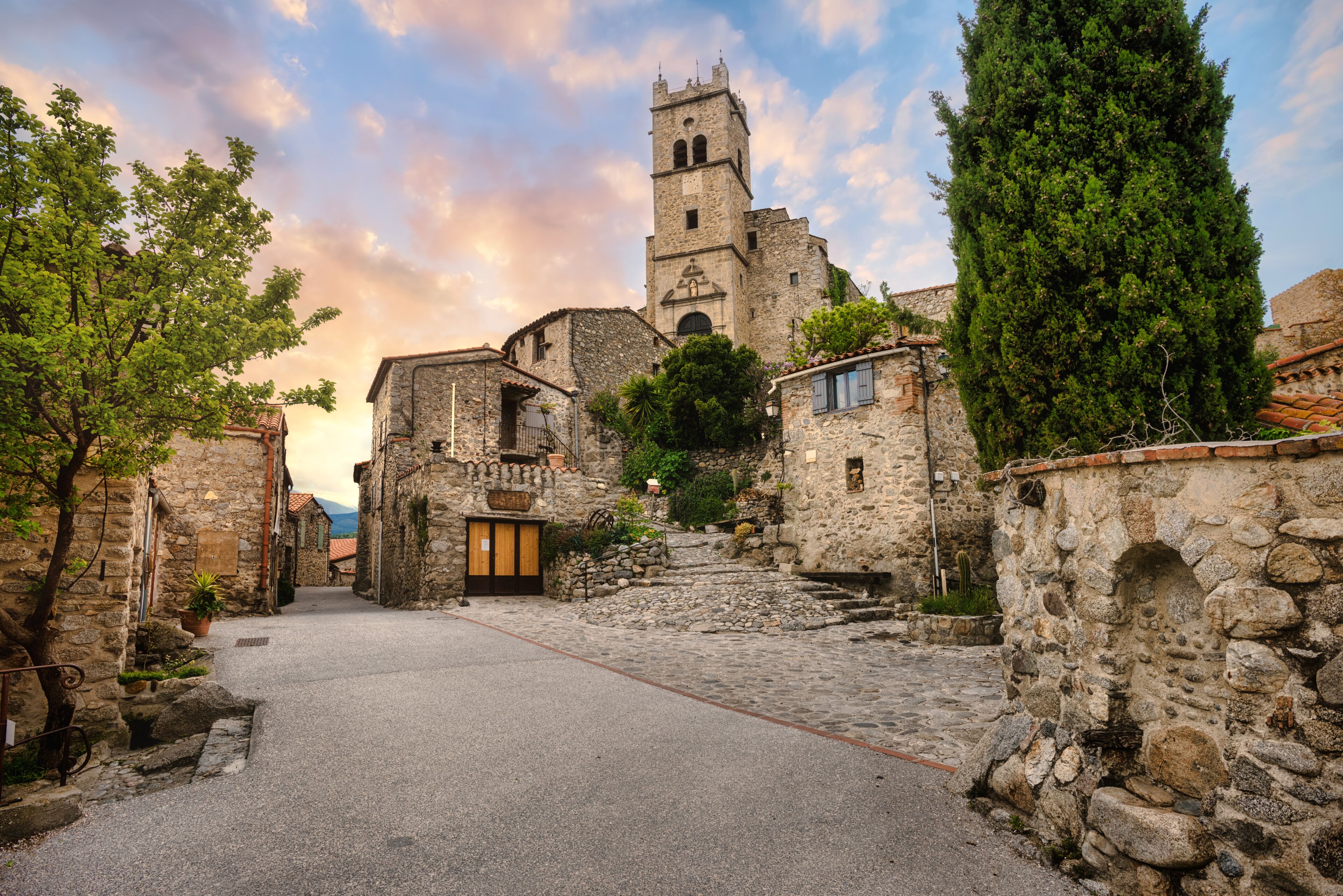 A cobblestone street leading to a historic stone building with a bell tower, surrounded by trees and stone walls.