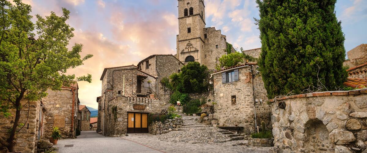 A cobblestone street leading to a historic stone building with a bell tower, surrounded by trees and stone walls.