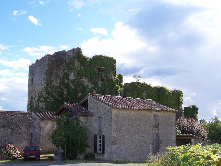 A stone building with a tiled roof, partially covered in ivy, and a parked car in front.