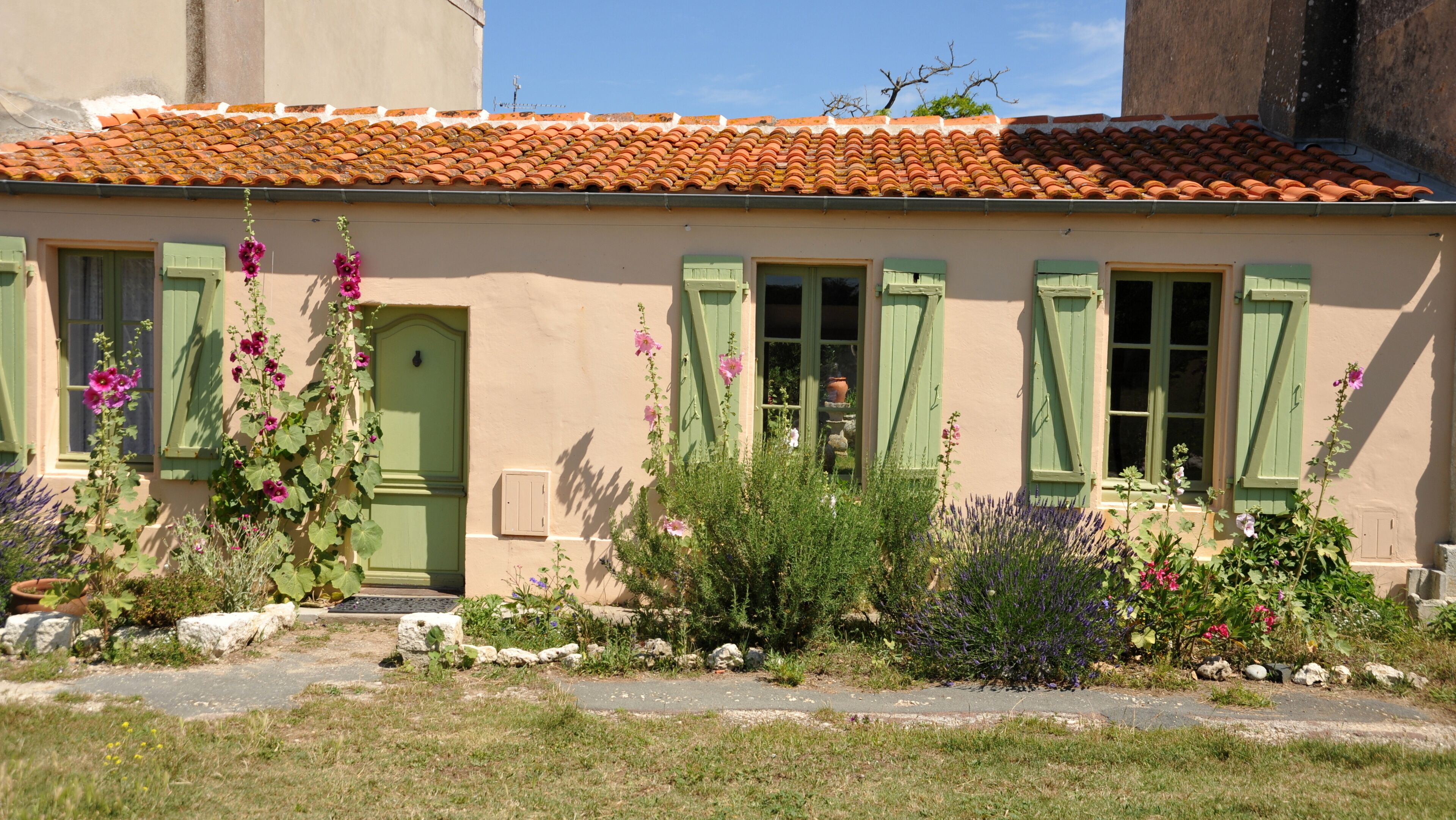 A quaint house with a tiled roof, green shutters, and a variety of flowers in the garden.