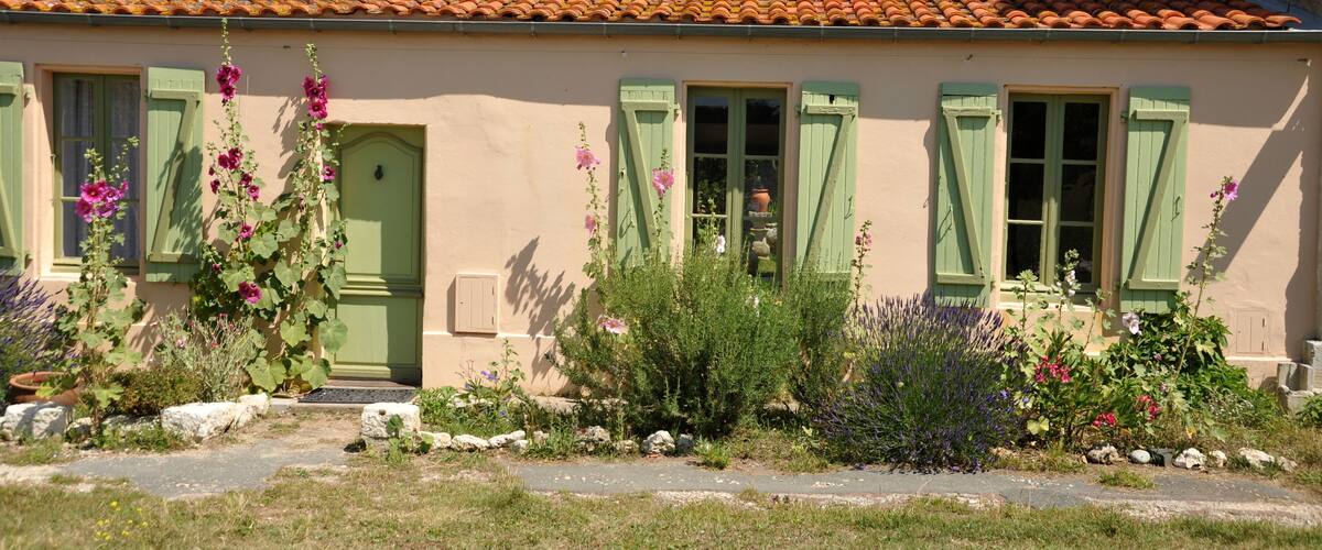 A quaint house with a tiled roof, green shutters, and a variety of flowers in the garden.