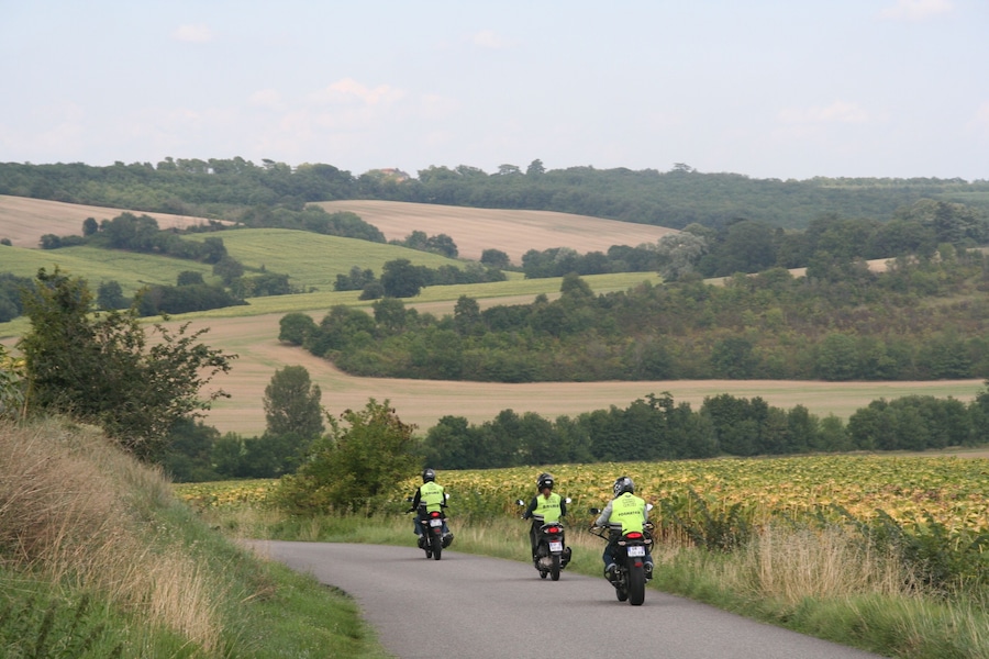 Tres motociclistas en una carretera rural, rodeados de colinas ondulantes y campos.