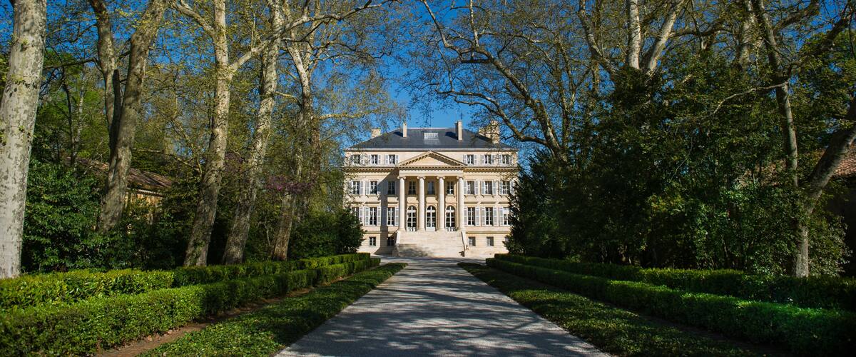 A grand mansion with a symmetrical facade, surrounded by a well-manicured garden and tall trees.