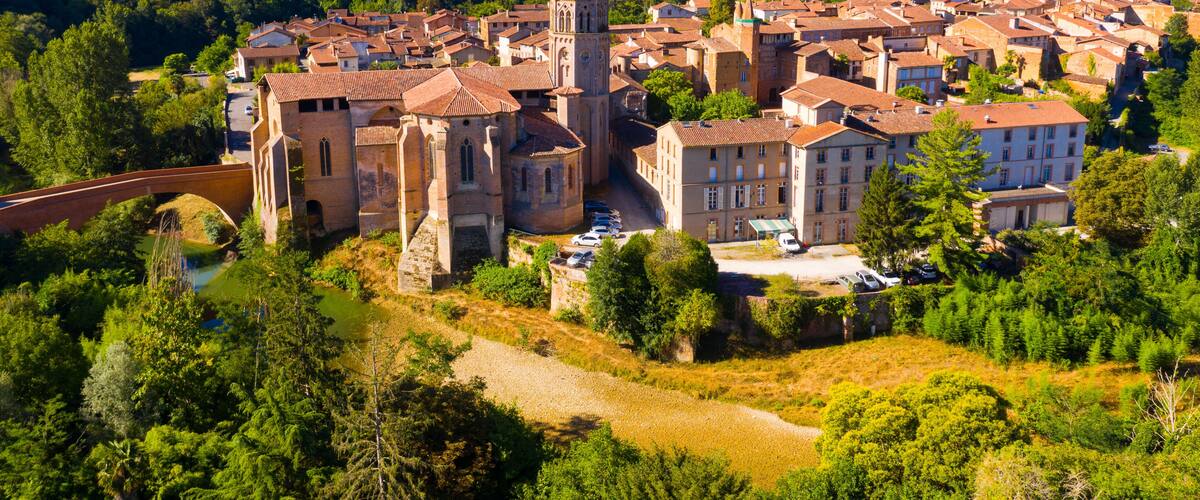 A historic town with a prominent church featuring a tall bell tower, surrounded by lush greenery and rolling hills in the background.