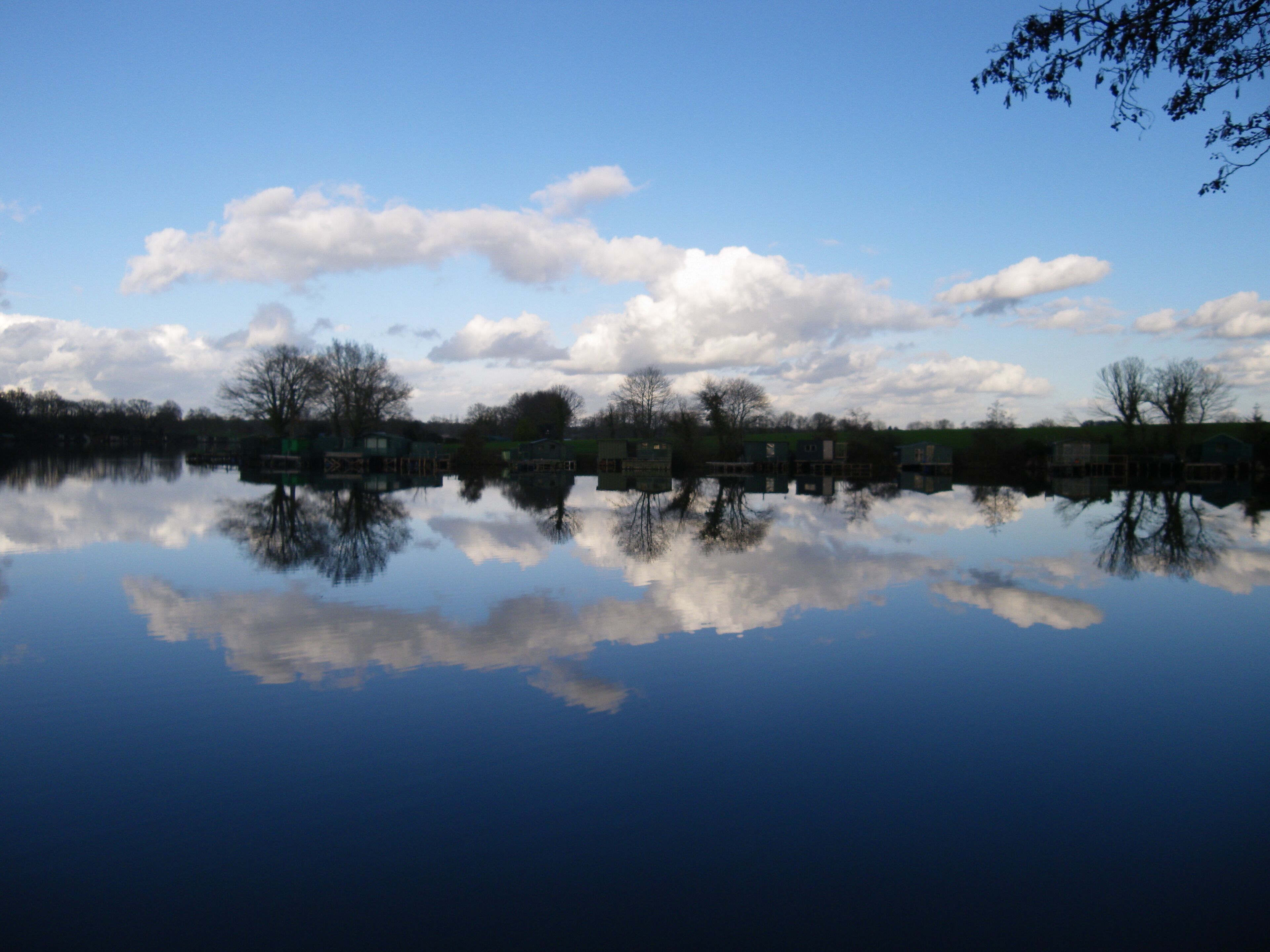 A calm lake with reflections of trees and buildings, clear sky, and scattered clouds.