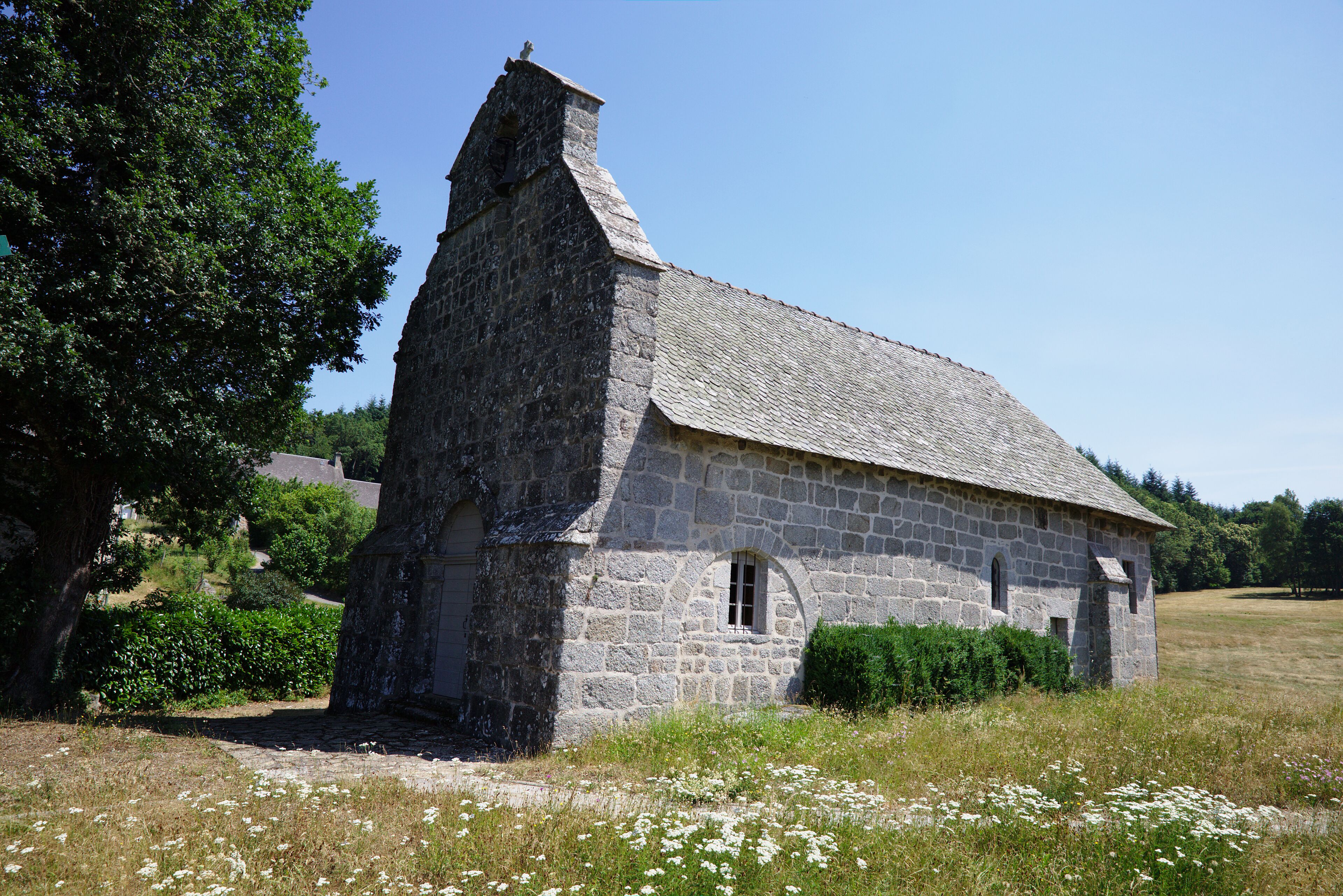 A stone church with a steep roof and a bell tower, surrounded by a grassy field and trees.