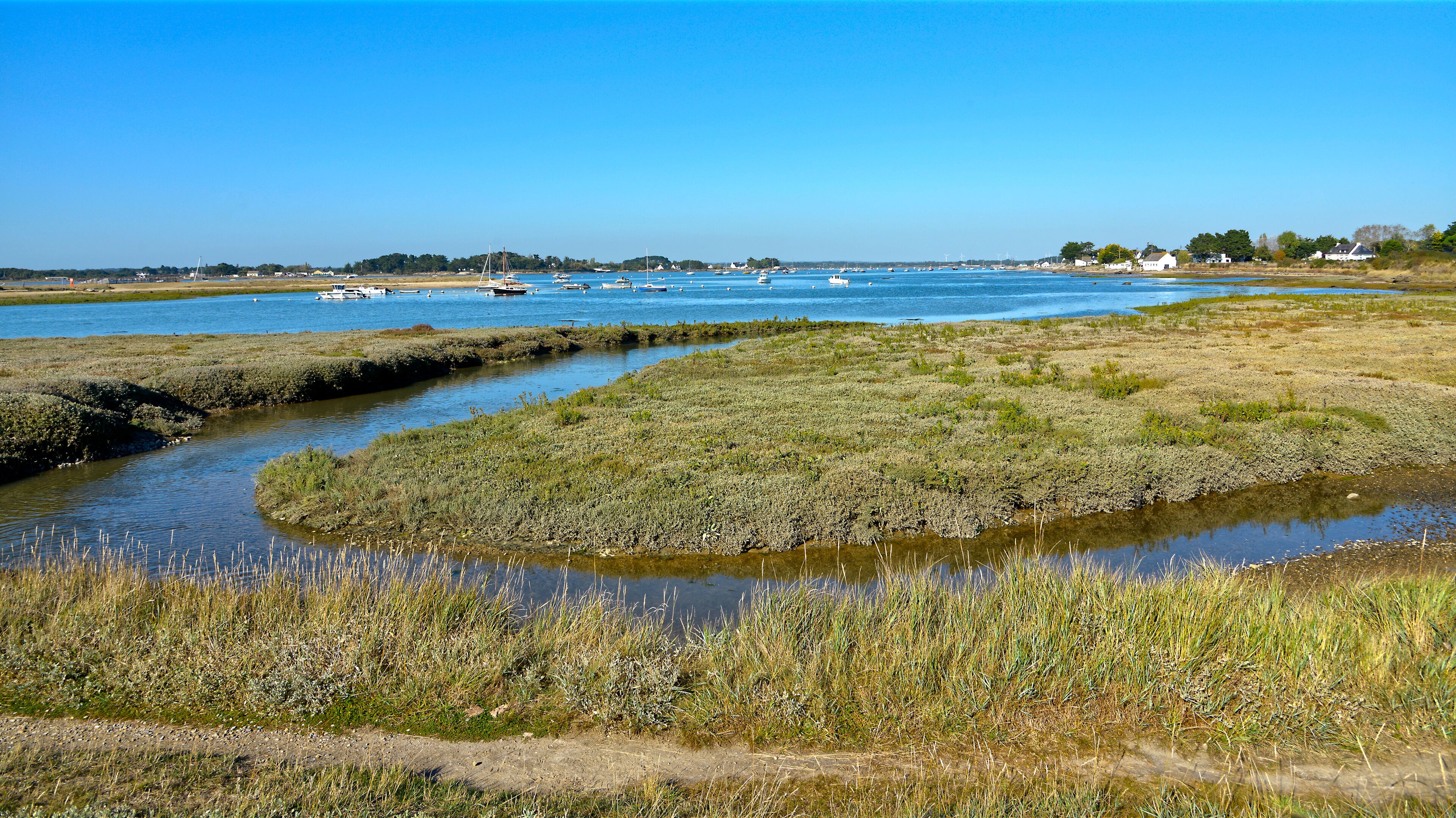 A coastal landscape with a river meandering through marshy terrain, boats on the water, and houses in the distance.