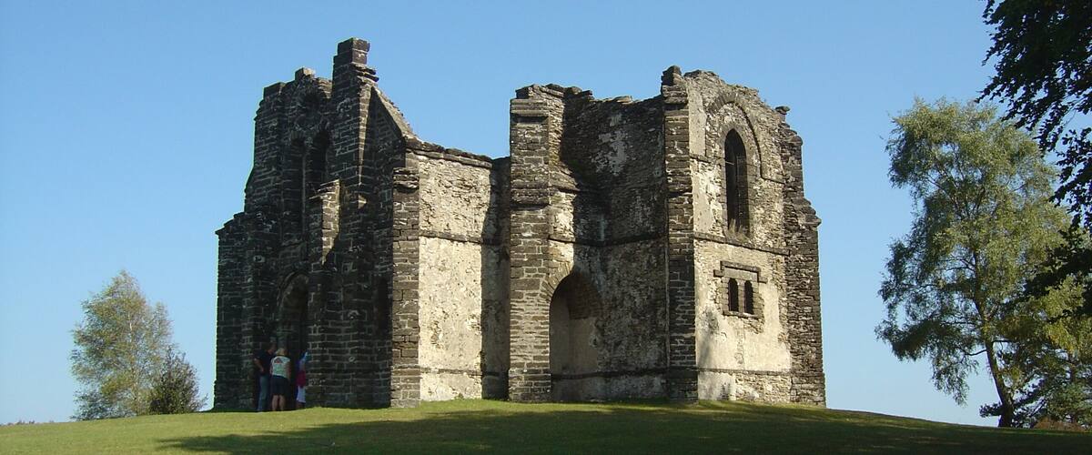 A historic stone building with arched windows and a tower, surrounded by grass and trees.