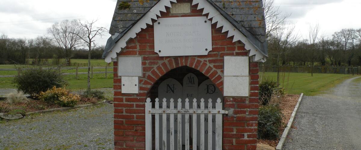A small brick chapel with a cross on top, a white fence, and a sign with inscriptions.