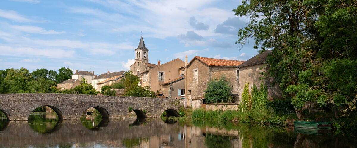 A stone bridge over a calm river, surrounded by lush greenery and historic buildings, including a church with a tall steeple.