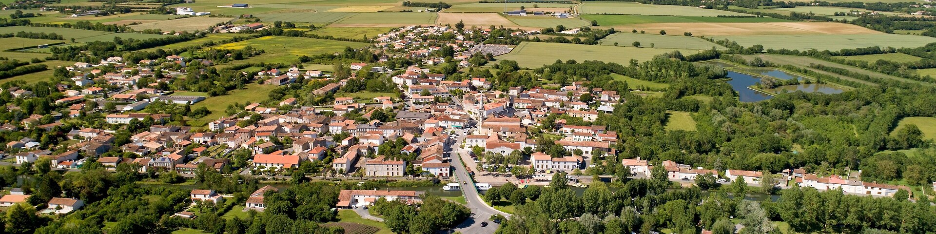 Vista aérea de uma aldeia rural com casas, campos e turbinas eólicas ao longe.