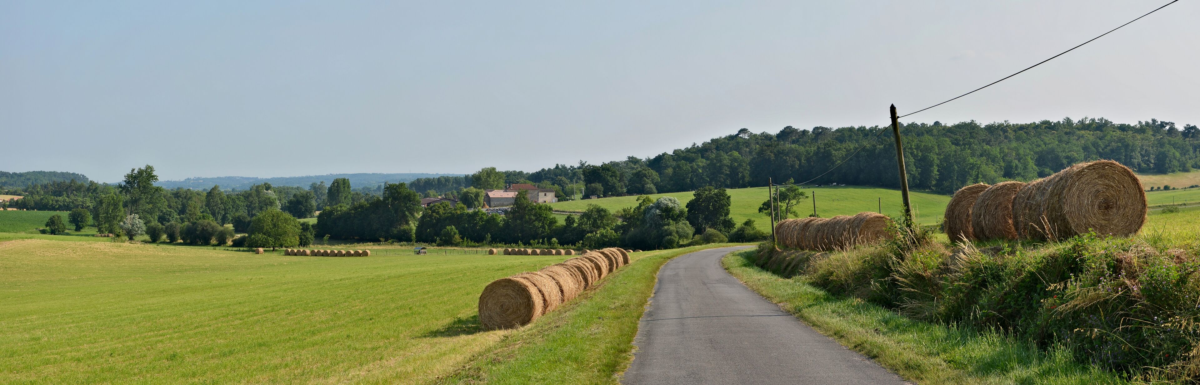 A rural road with hay bales on the side and a house in the distance.