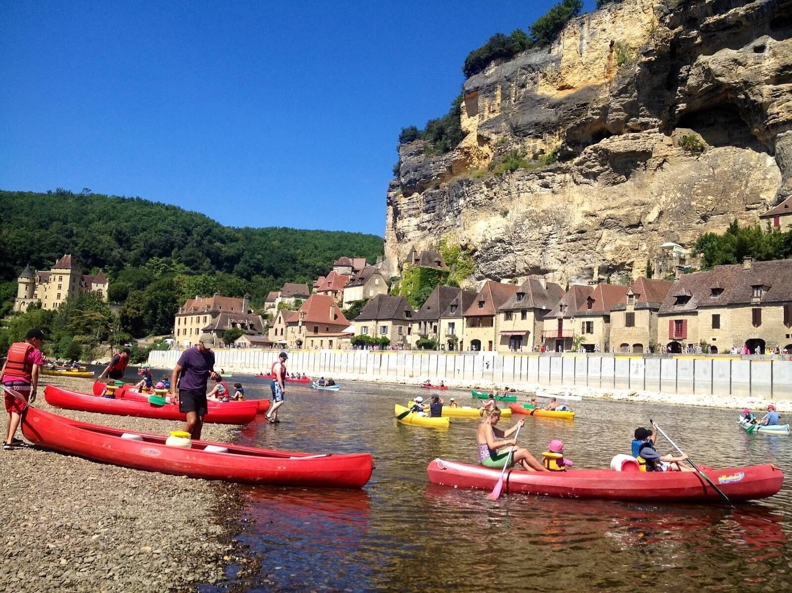 A group of people in red canoes on a calm river, with a village and rocky cliffs in the background.