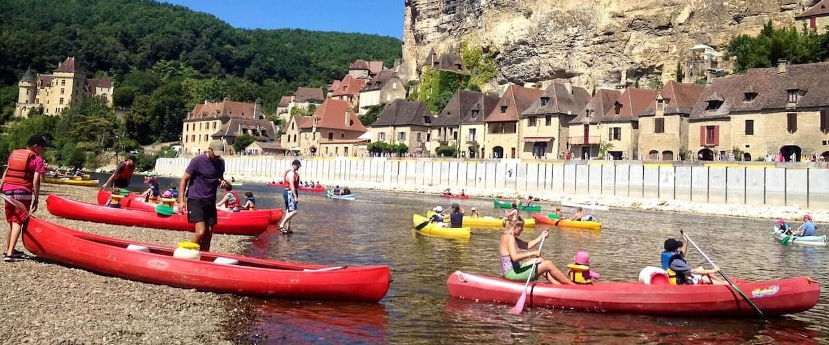 A group of people in red canoes on a calm river, with a village and rocky cliffs in the background.