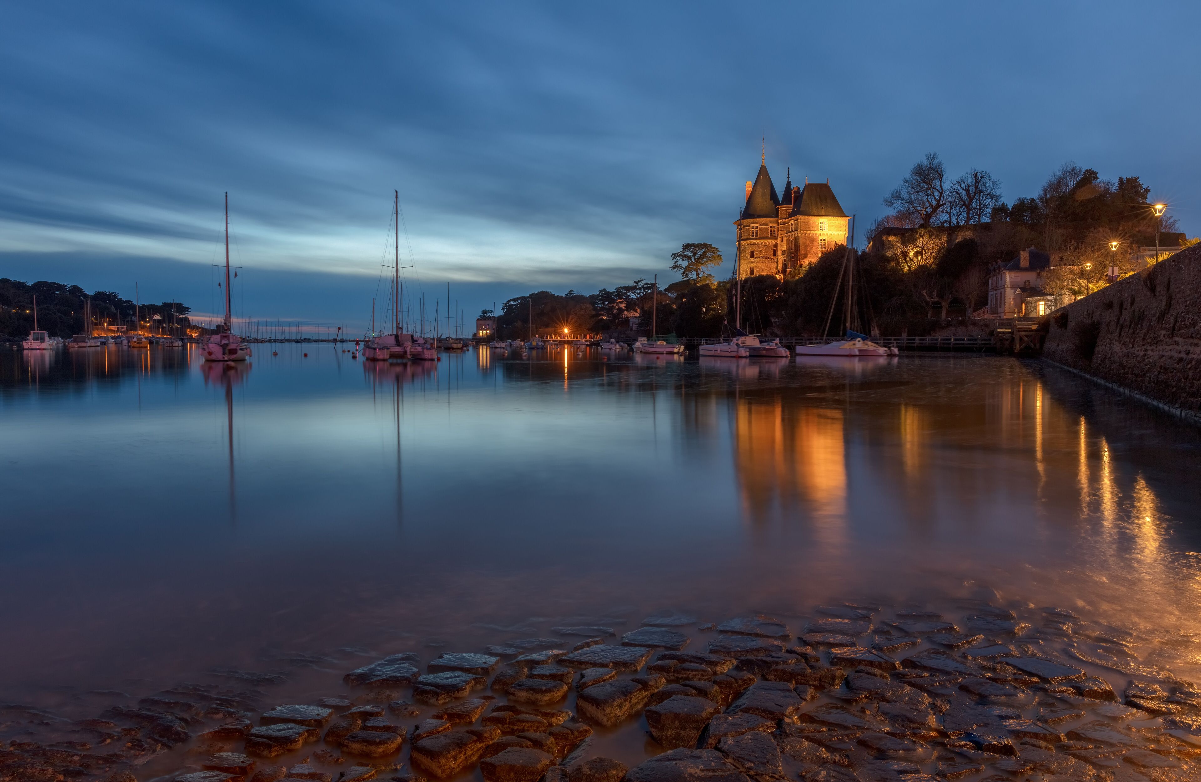 A calm harbor at dusk with boats, a castle, and cobblestone walkways.