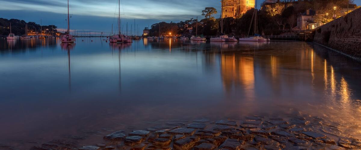 A calm harbor at dusk with boats, a castle, and cobblestone walkways.