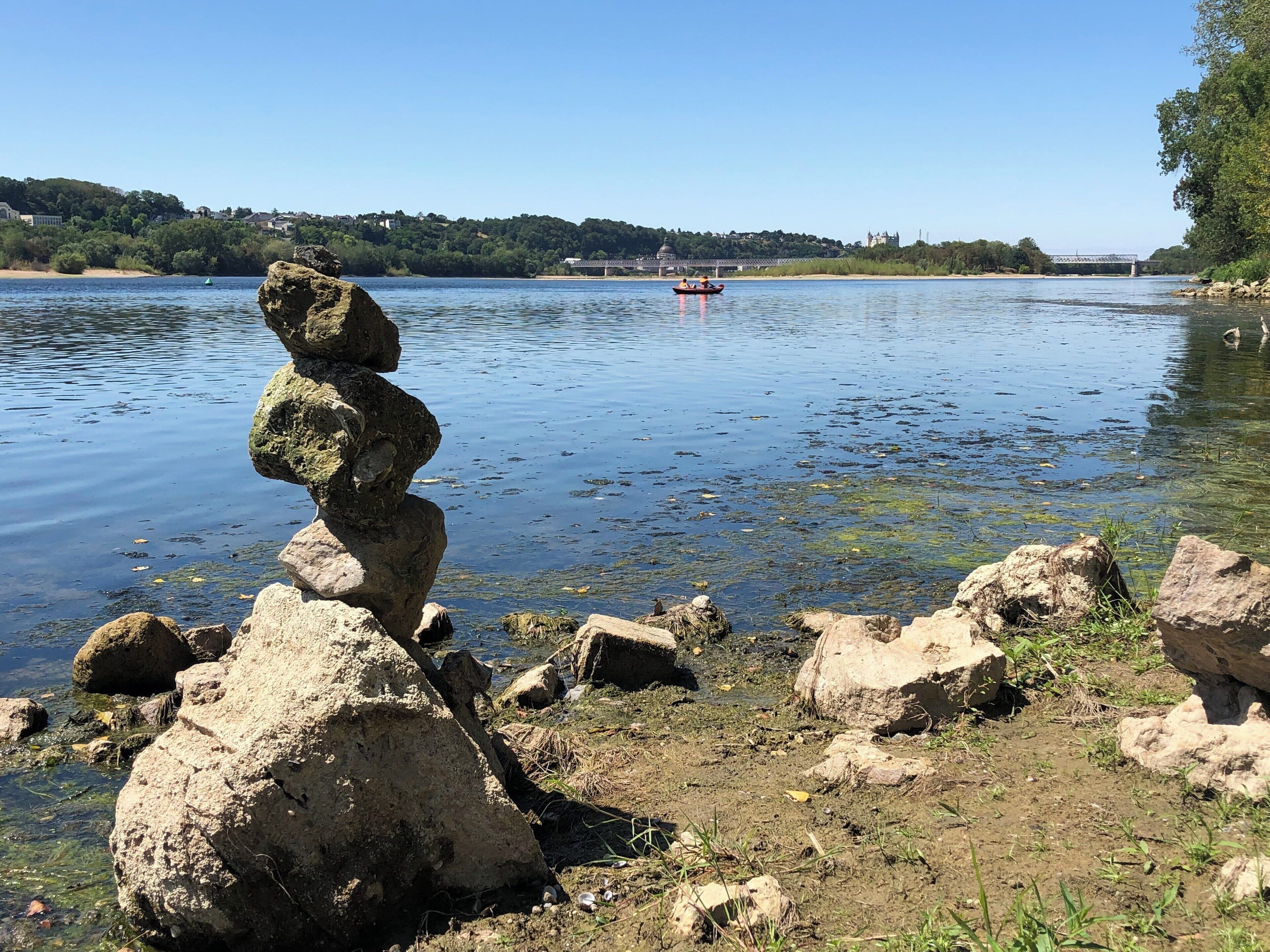 A calm lake with a stack of rocks in the foreground, a bridge in the background, and a castle on a hill.