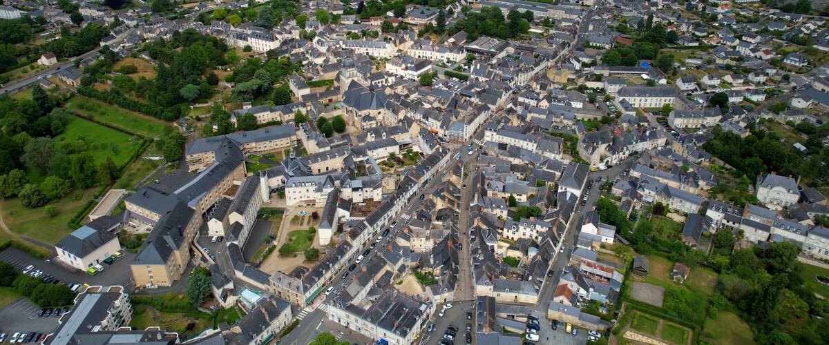 An aerial view of a densely built town with historic architecture, surrounded by green fields and a large stadium.