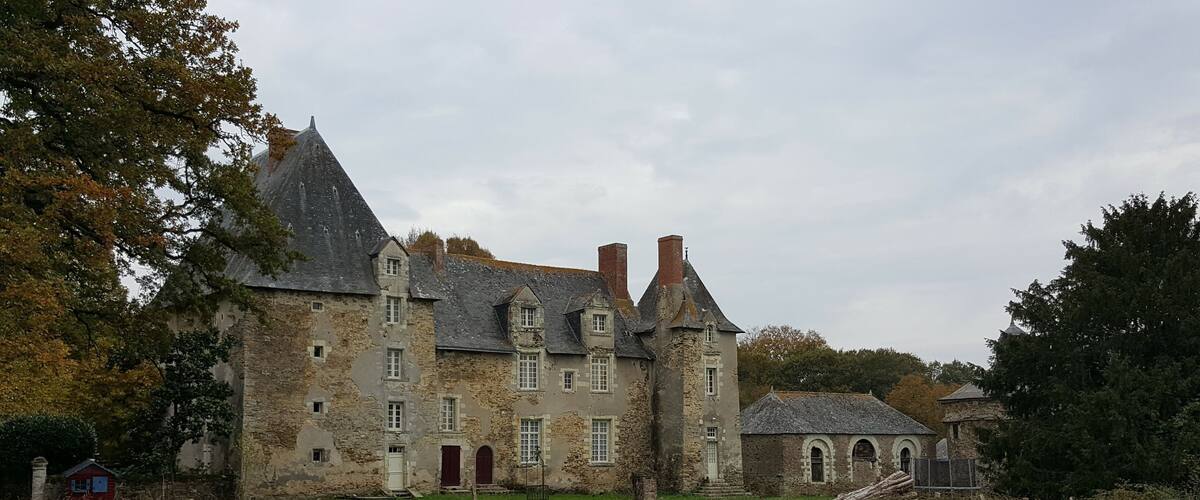 A historic stone building with multiple chimneys and arched windows, surrounded by trees and a grassy field.
