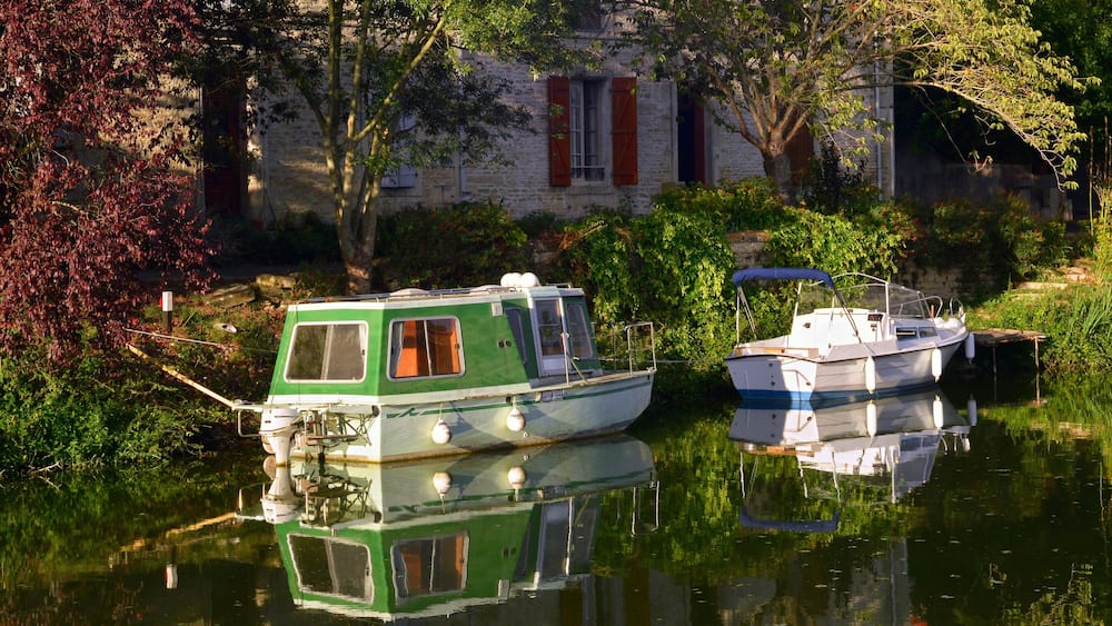 A green and white houseboat docked near a stone building with red shutters.