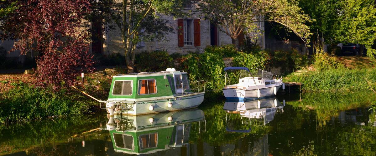 A green and white houseboat docked near a stone building with red shutters.