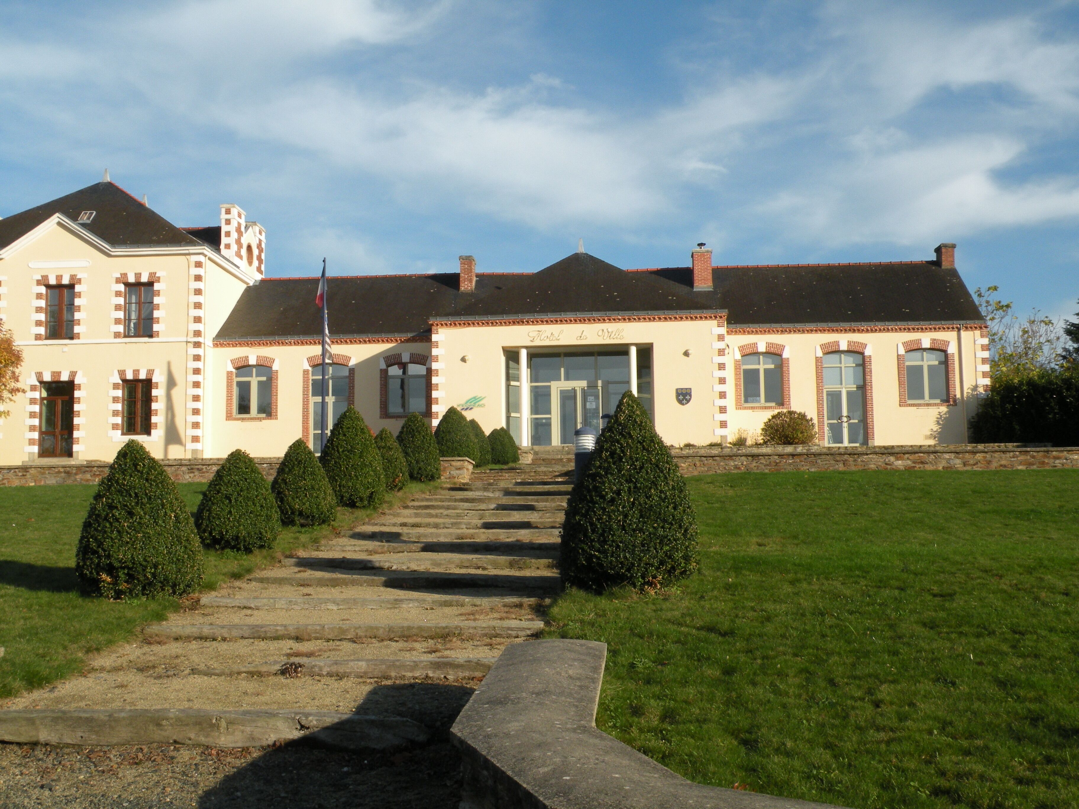 A large, historic building with a symmetrical facade, a central entrance, and a well-maintained lawn with trimmed bushes.