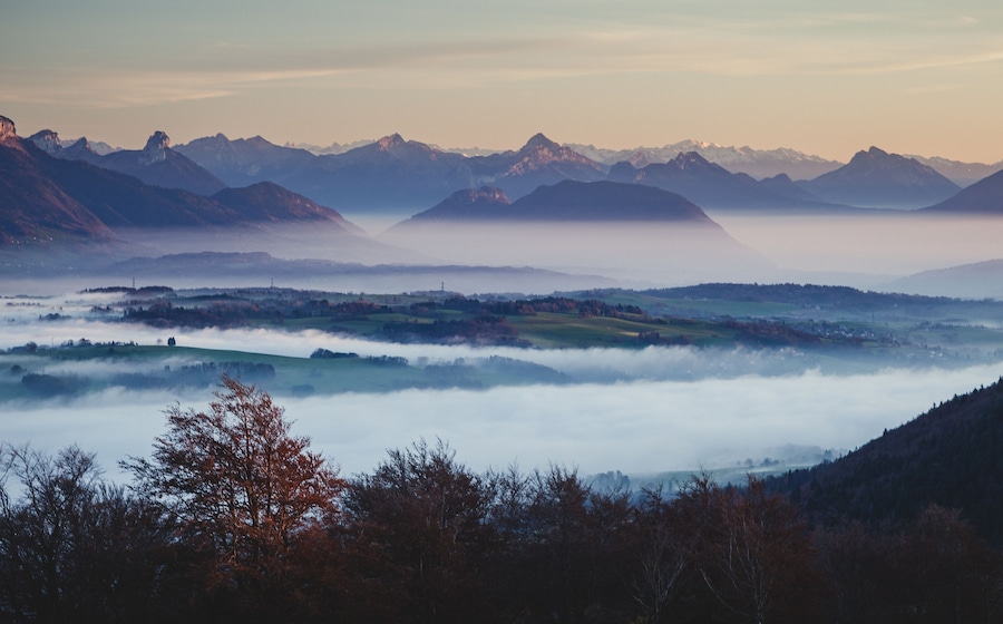A mountainous landscape with a valley shrouded in mist, distant peaks, and a foreground of trees.