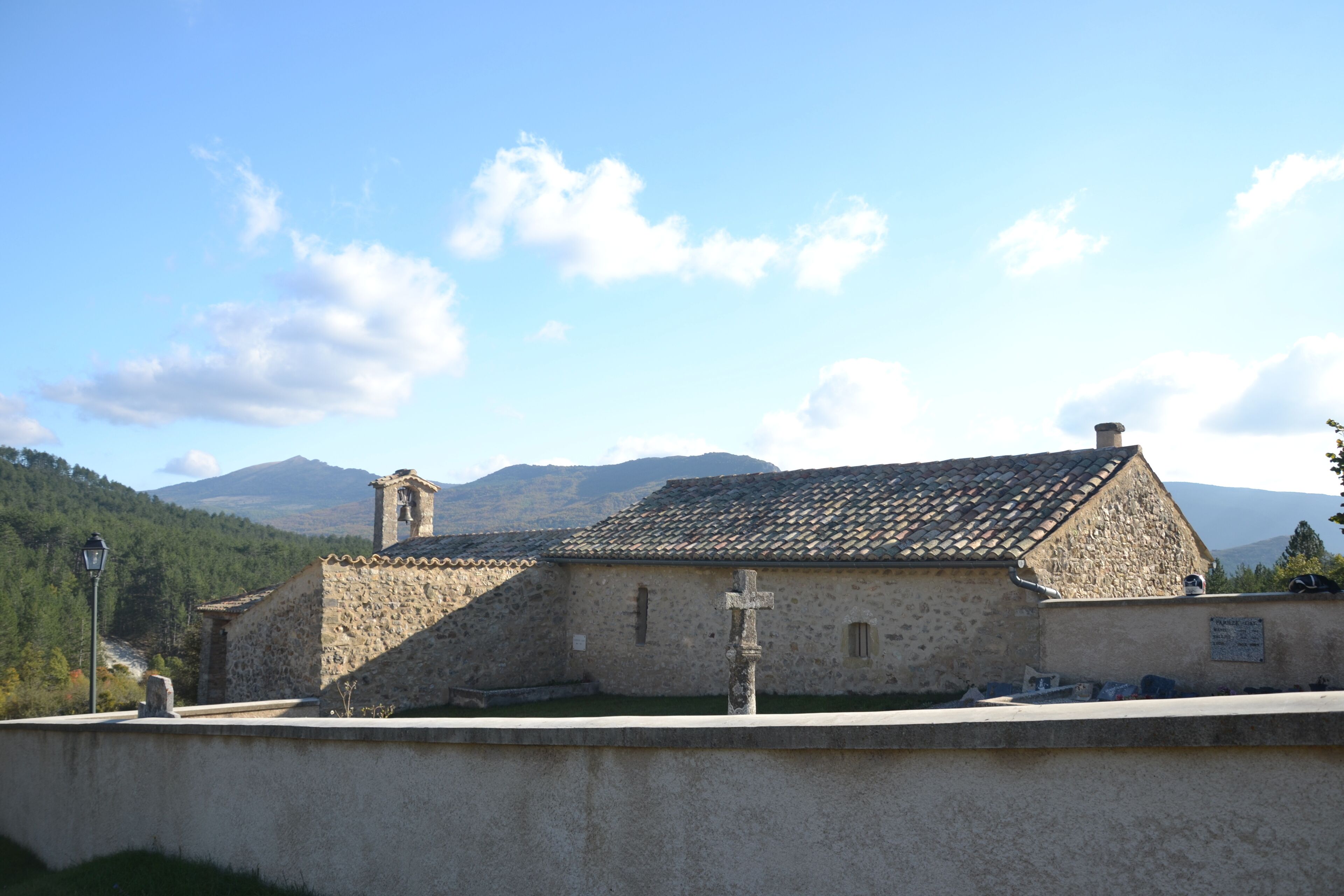 A stone church with a bell tower and a cross in front, set against a mountainous backdrop.