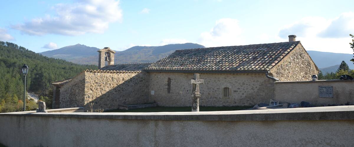 A stone church with a bell tower and a cross in front, set against a mountainous backdrop.