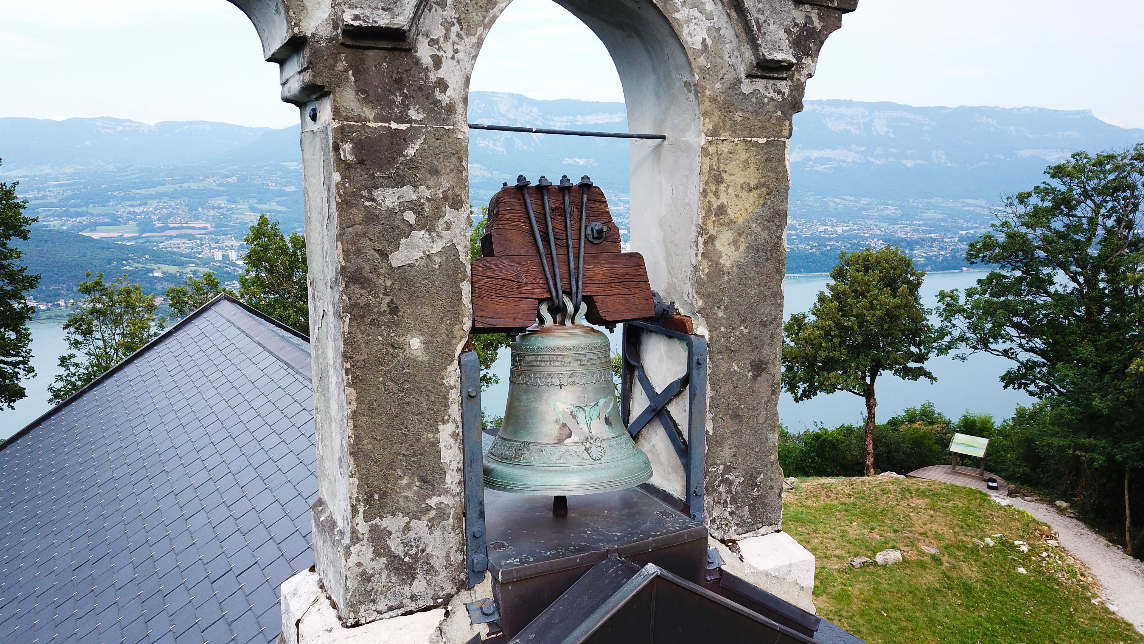 A bell tower with a bell, surrounded by a stone archway, overlooking a lake and mountains.