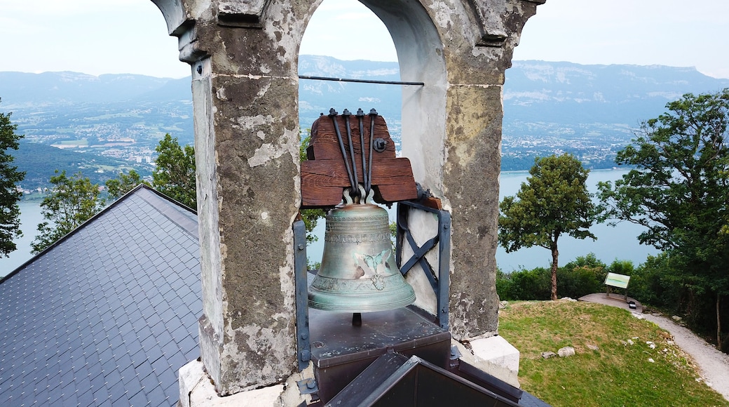 A bell tower with a bell, surrounded by a stone archway, overlooking a lake and mountains.