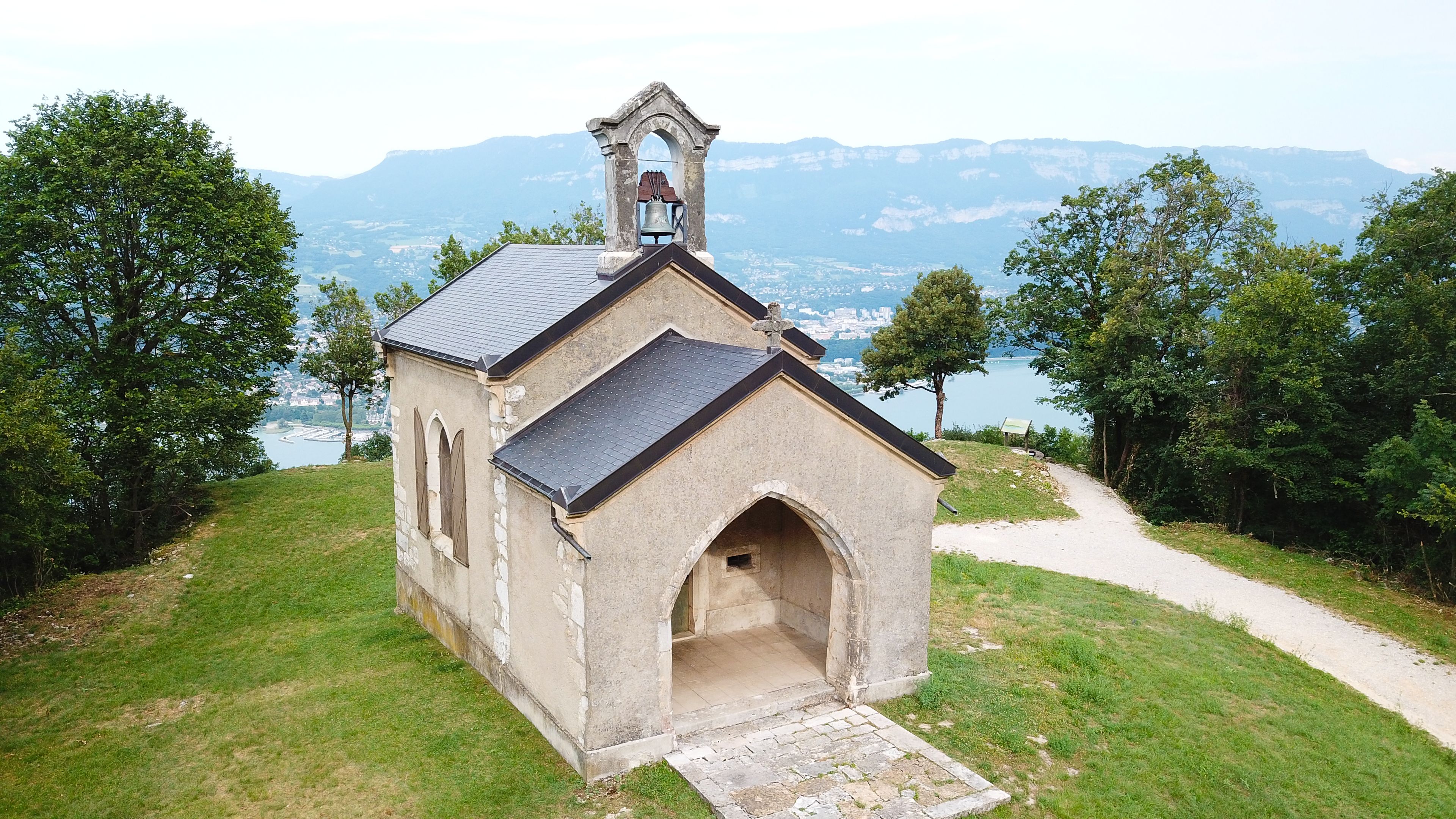 A small church with a bell tower, surrounded by trees and a lake with mountains in the background.