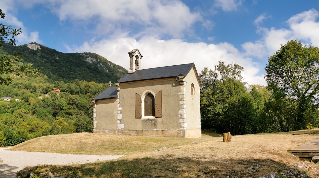 A small stone church with a bell tower, surrounded by lush greenery and mountains in the background.
