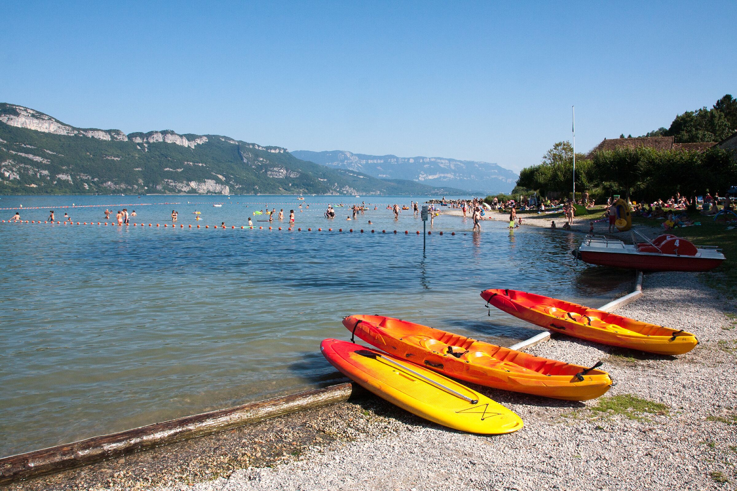 A lake with kayaks on the shore, people swimming, and mountains in the background.