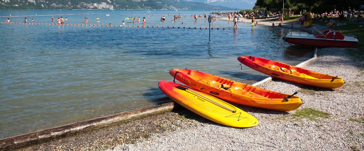 A lake with kayaks on the shore, people swimming, and mountains in the background.