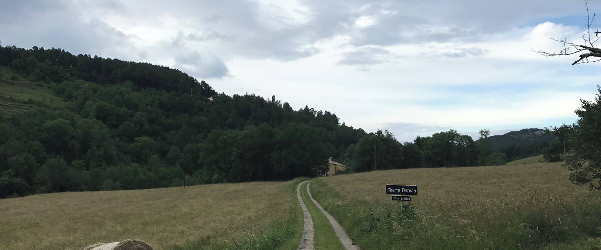 A gravel path leading through a rural landscape with a hay bale on the left and a sign for 'Champ Terreau' in the distance.