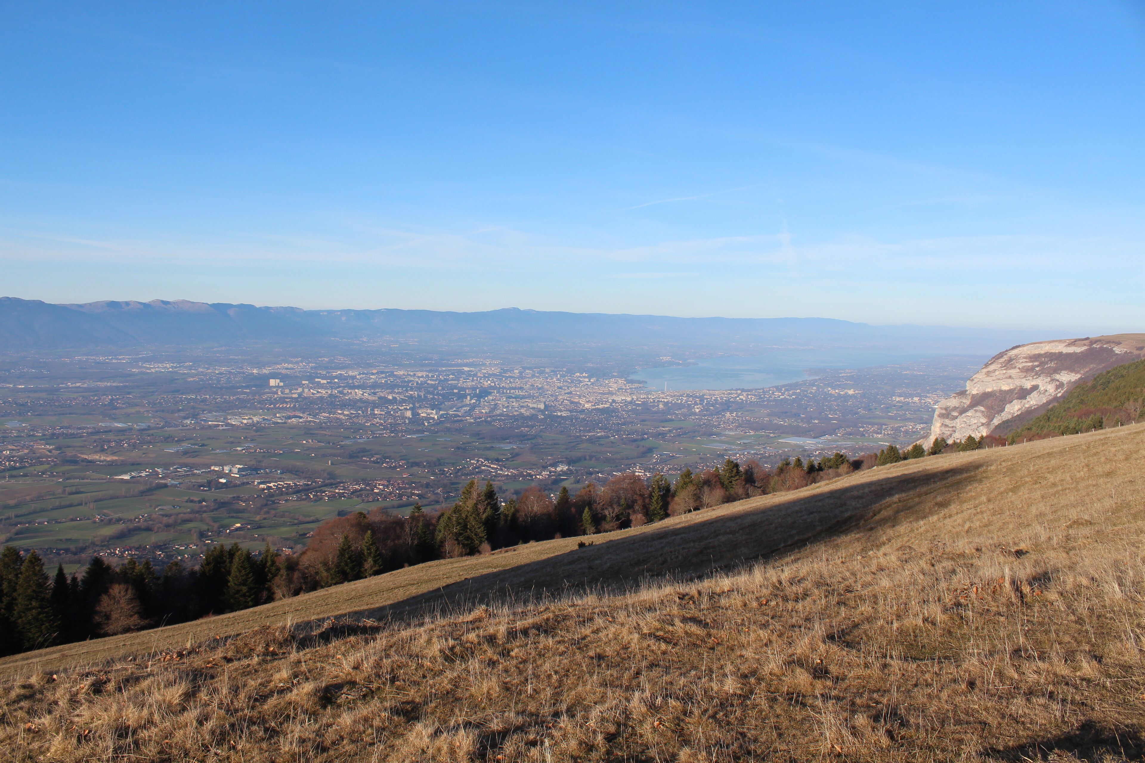 A panoramic view of a valley with a distant city, a hill with sparse vegetation, and a clear blue sky.