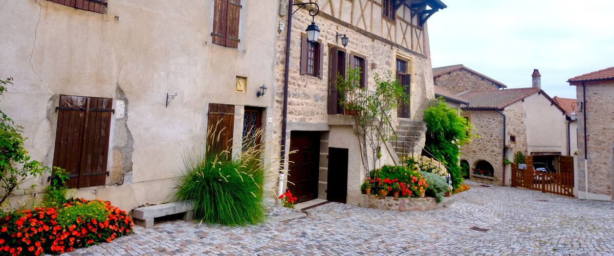 A cobblestone courtyard with a half-timbered building, wooden shutters, and flowering plants.