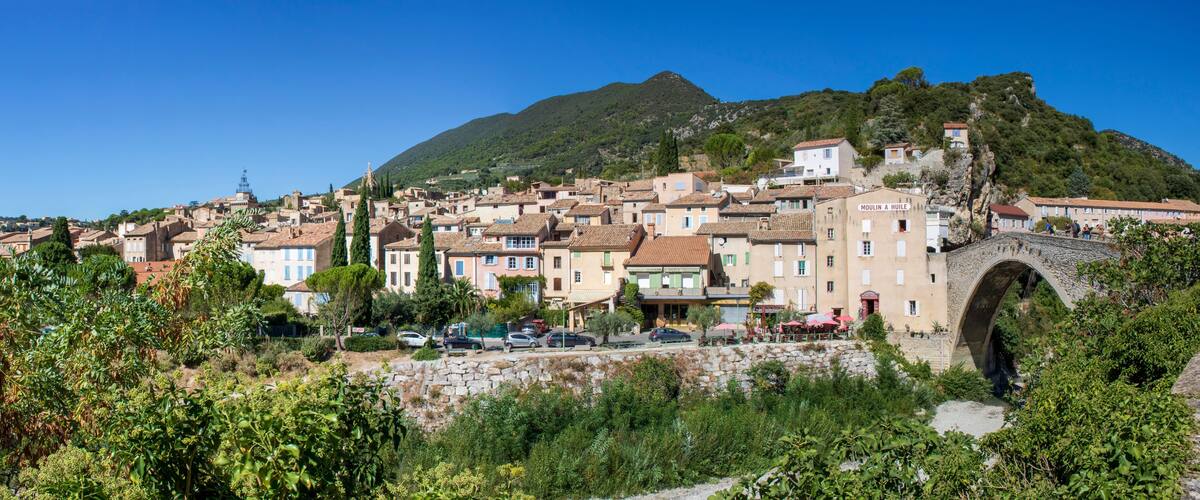 A coastal town with terracotta-roofed buildings, a stone bridge, and a mountain in the background.