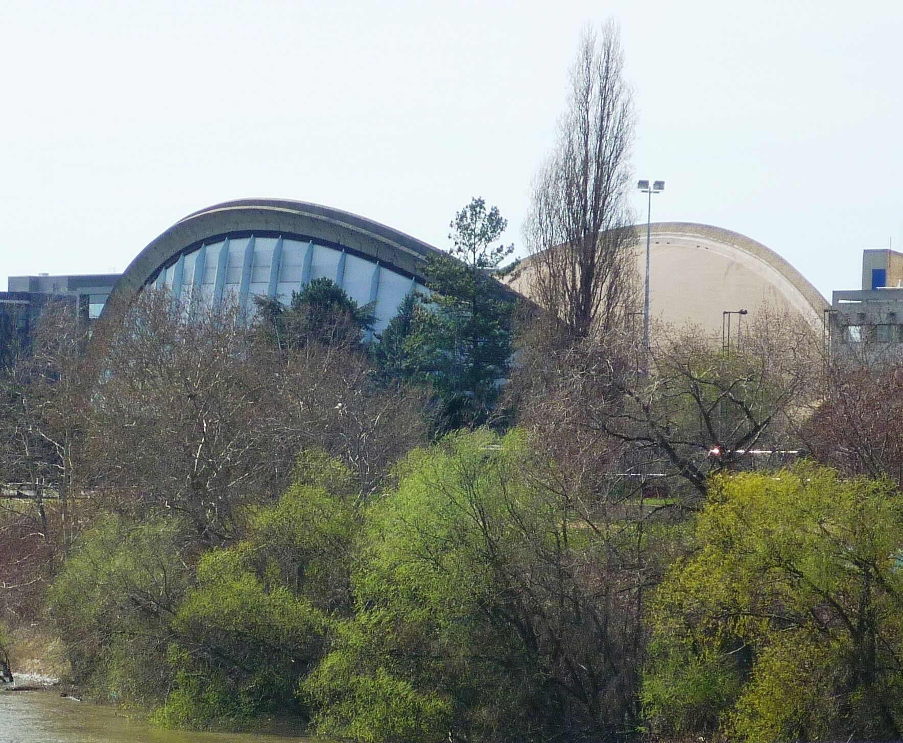 A modern building with a curved glass facade, surrounded by trees and a large dome structure in the background.