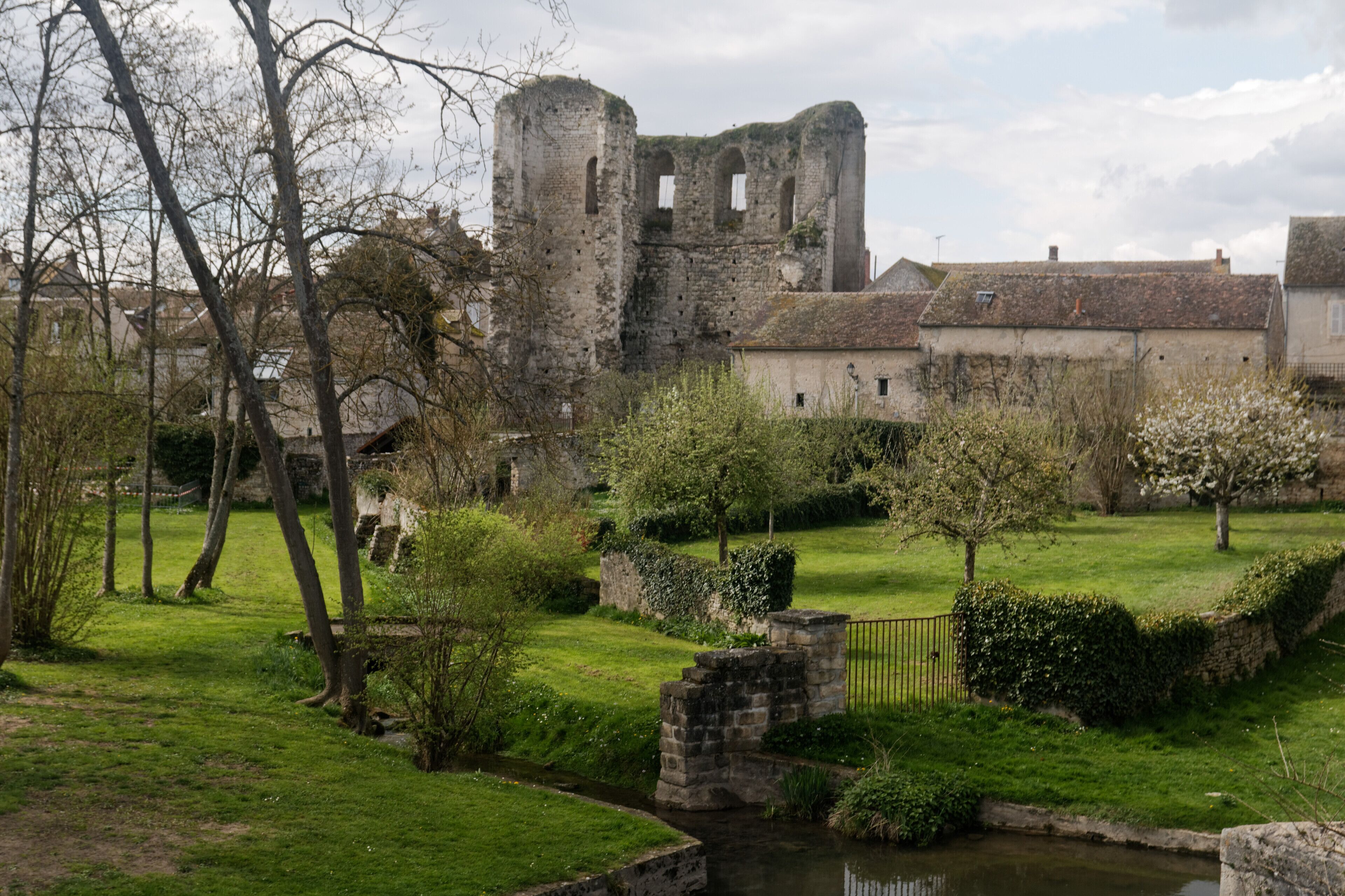 A historic stone castle with ruins, a small stream, and a well-maintained garden with trees and a hedge.